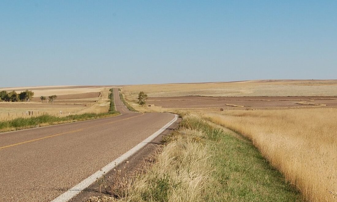 A two-lane highway curving across golden grassland on Montana's Hi-Line under a clear sky
