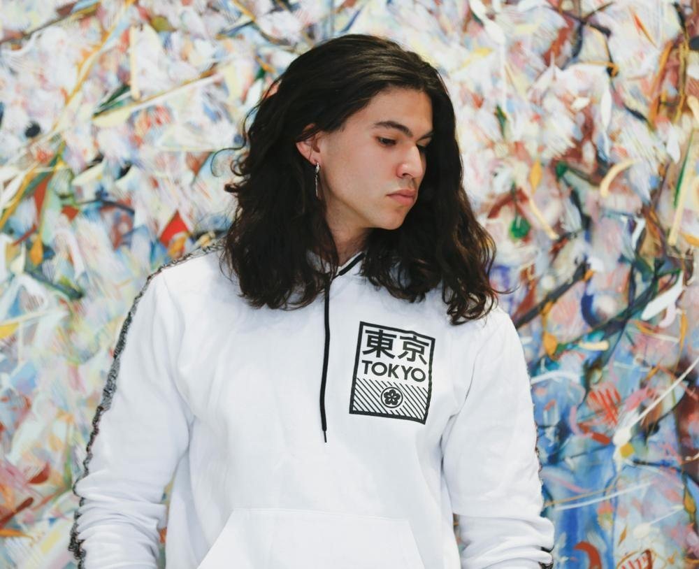 Wiingashk Courchane in a white hoodie looking to the side in front of a colorful abstract painting